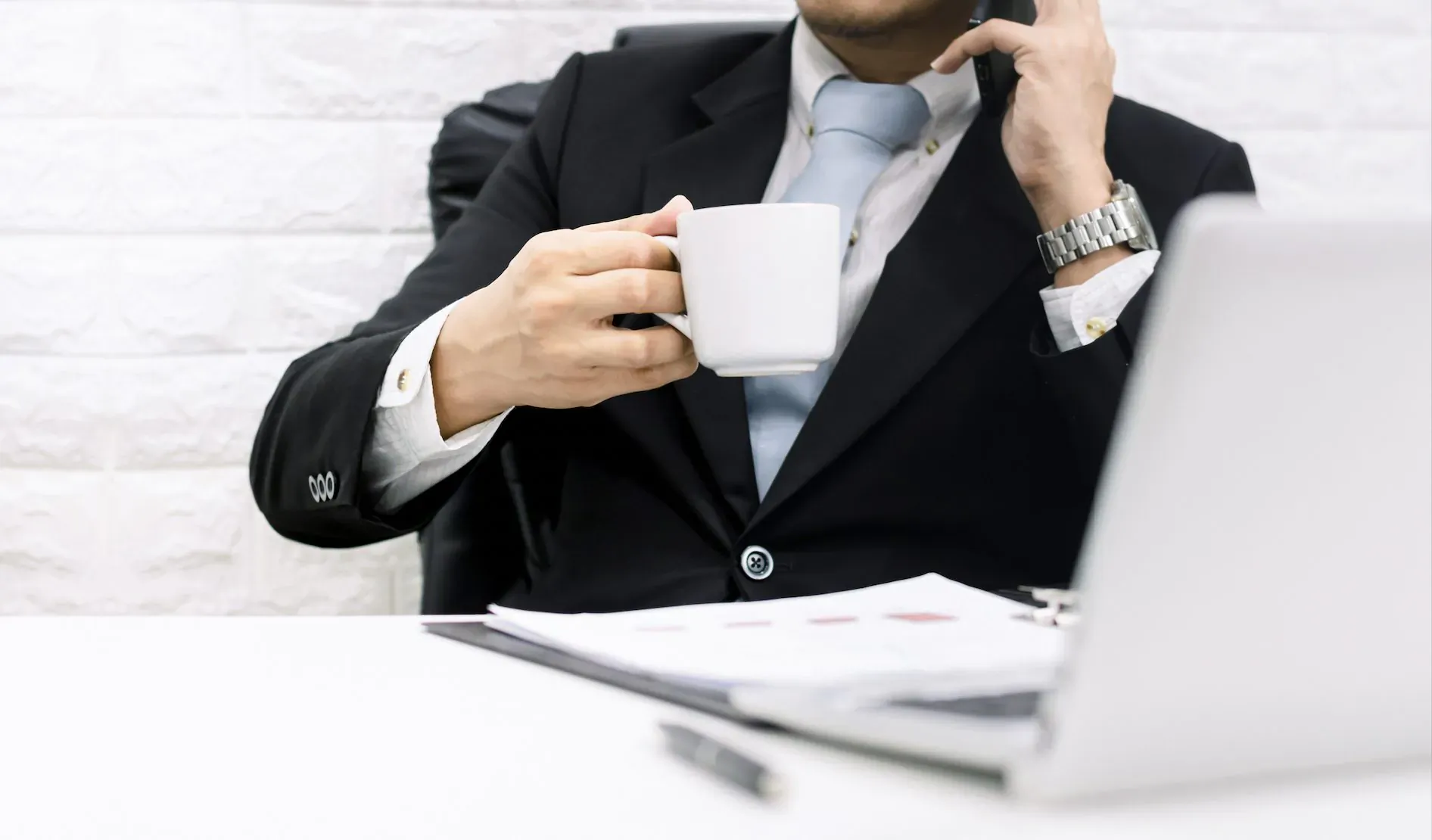 Businessman in suit sips white coffee mug while on phone call at desk with laptop and papers