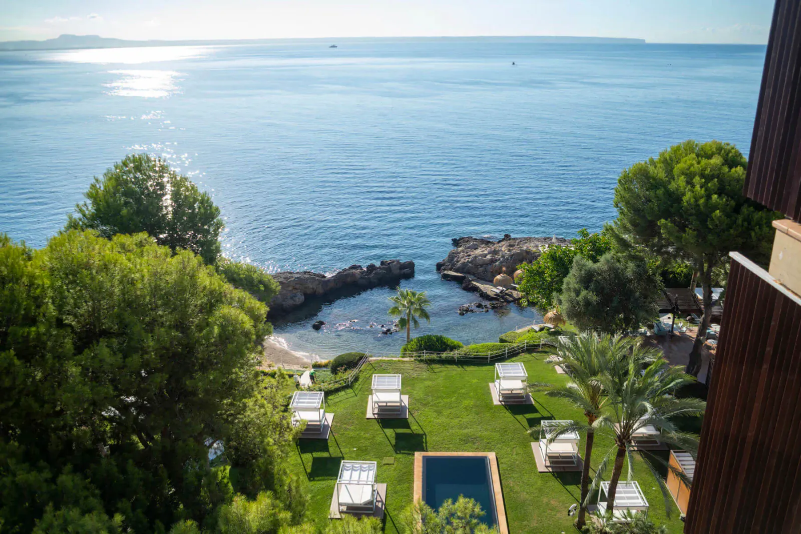 Aerial view of luxury hotel terrace with lounge chairs, pool, palm trees, overlooking rocky cove and Mediterranean Sea at sunset.