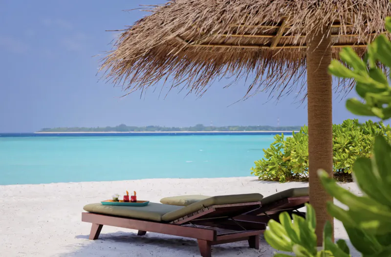 Luxury beach lounger with tray of red drinks under thatched umbrella, turquoise ocean and white sand at Amari Raaya Maldives