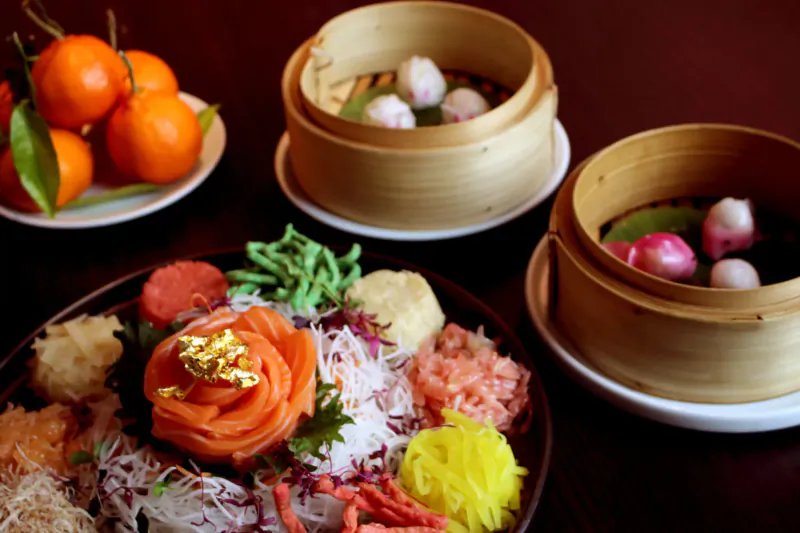 Vibrant yu sheng salad with gold salmon rose, oranges, bamboo steamers of dumplings on wooden table