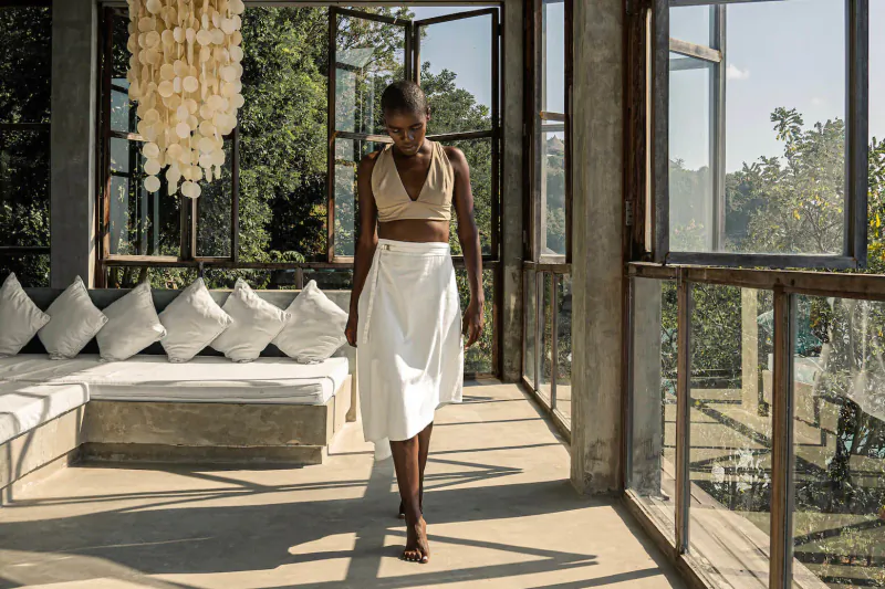 Black woman in beige bralette and white skirt walks on sunlit resort veranda with shell chandelier and lush garden view