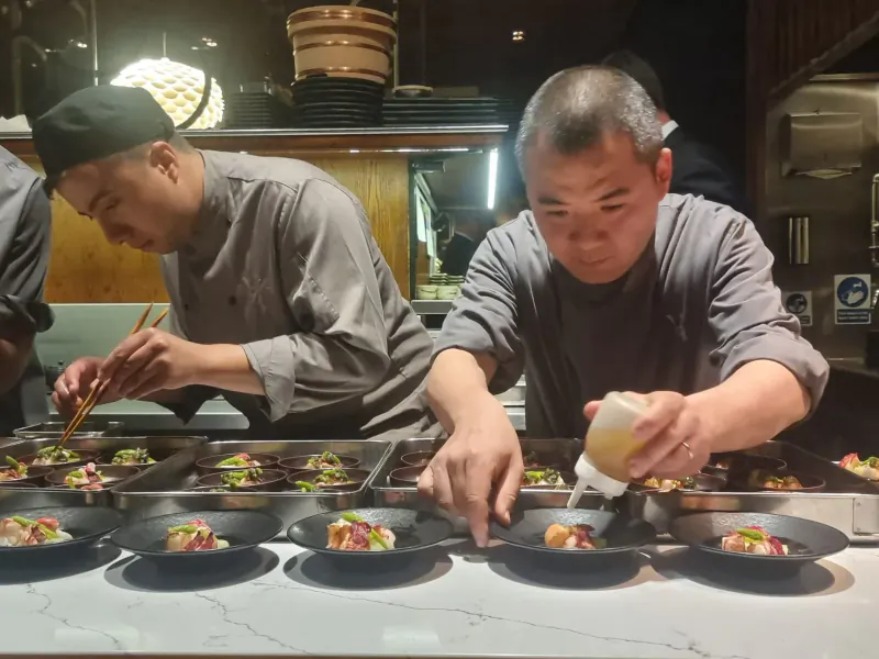 Chefs in UMU kitchen plating colorful dishes: one drizzling sauce, another using chopsticks on plates.