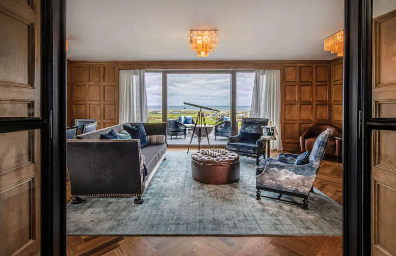 Elegant hotel living room with gray sofas, armchairs, drum stool, telescope by window overlooking green fields.