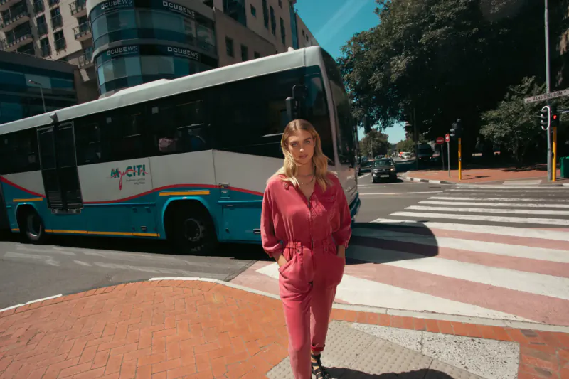 Blonde woman in pink jumpsuit stands stylishly at urban crosswalk beside MyCiTi bus, Cape Town streetscape.