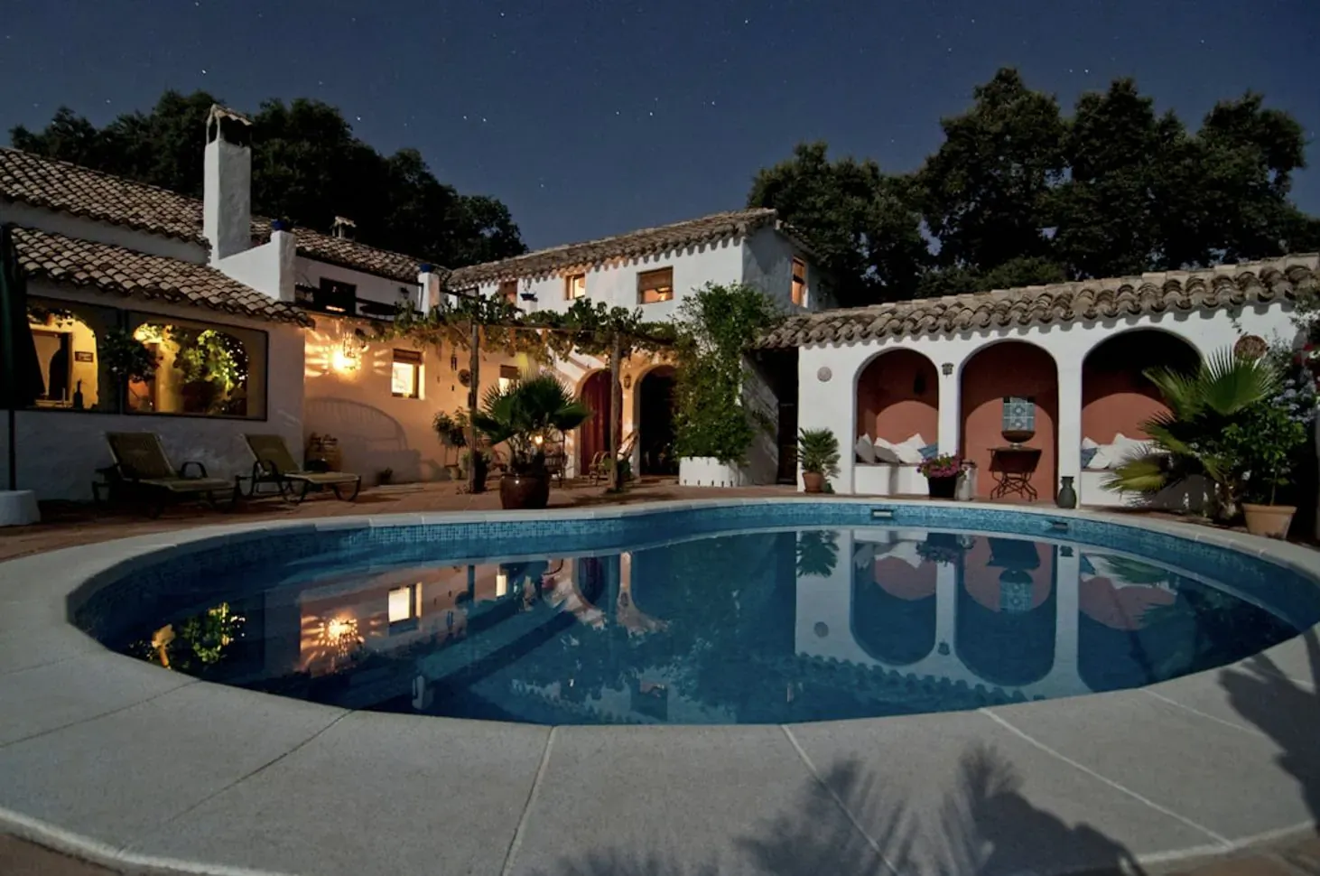 Night view of luxury Spanish-style villa with lit pool, patio furniture, plants, and starry sky.