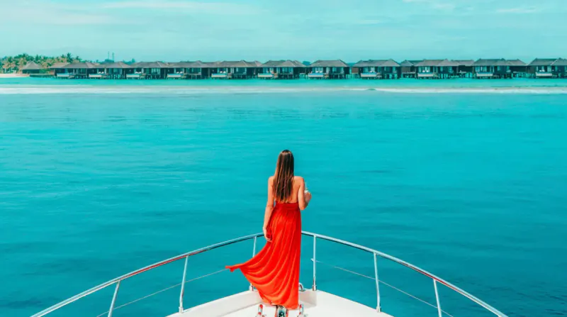 Woman in flowing red dress stands at the bow of a luxury yacht facing turquoise sea and overwater bungalows