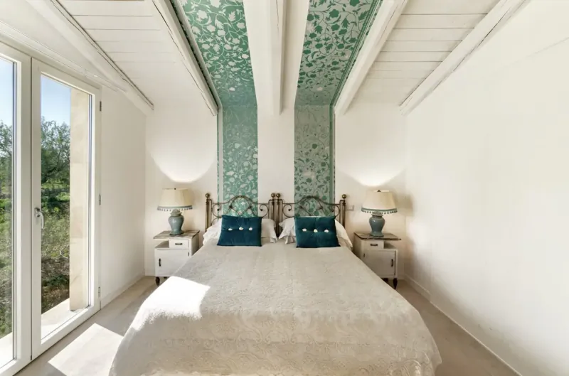 Elegant bedroom in Sicilian country house with teal leaf-patterned walls, white king bed, lamps, and garden view.