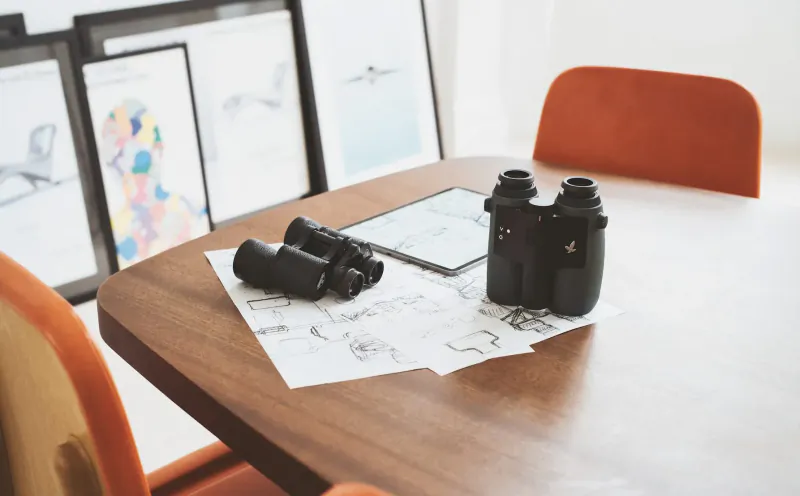 Smart binoculars by Swarovski Optik on wooden table with sketches, orange chair, and art frames in background.