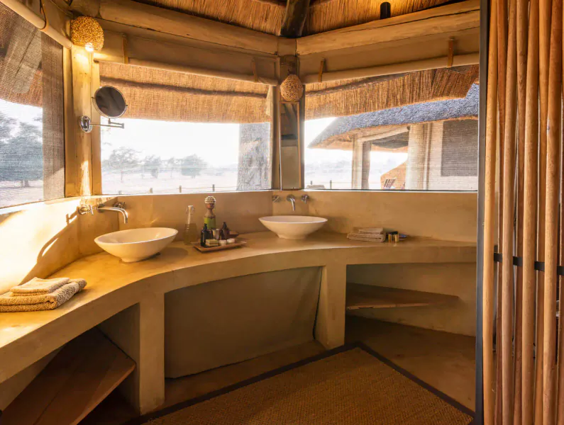 Rustic thatched Lion Camp bathroom with curved wooden vanity, white sinks, towels, and savanna view