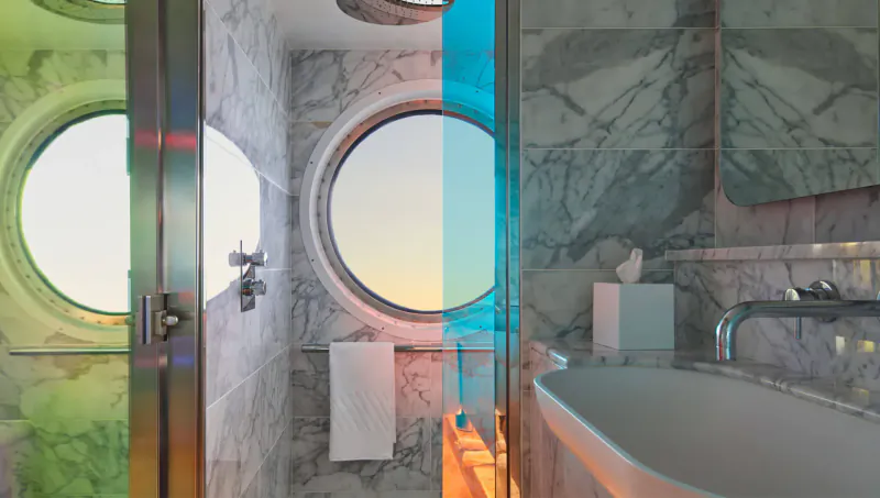 Luxurious adults-only cruise bathroom with marble walls, round porthole sunset view, colorful lighting, sink, and towels.