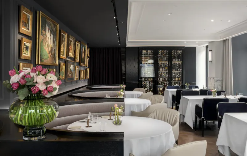 Elegant Vienna gallery restaurant with curved tables, white linens, pink roses in vase, and framed art on dark walls.