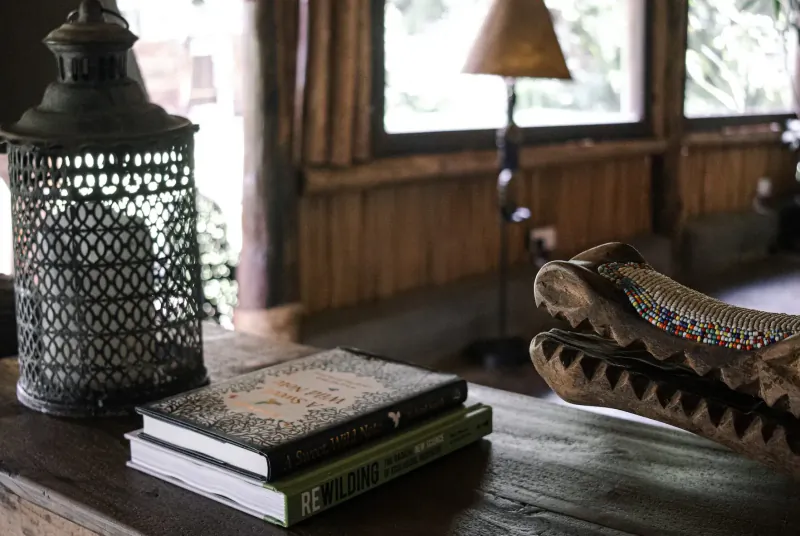 Rustic wooden table with stacked books, ornate metal lantern, beaded crocodile skull, in Kenyan wild house interior.