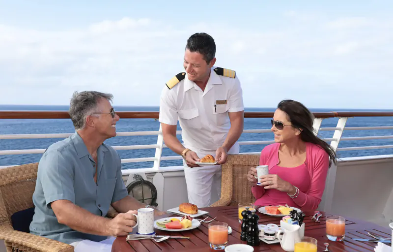On luxury yacht deck, uniformed server offers breakfast plates to smiling older couple at ocean-view table with fruits, coffee, juice.