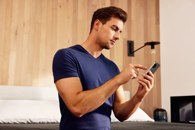 Fit man in navy V-neck T-shirt taps smartphone screen in modern bedroom with wood wall and bed.