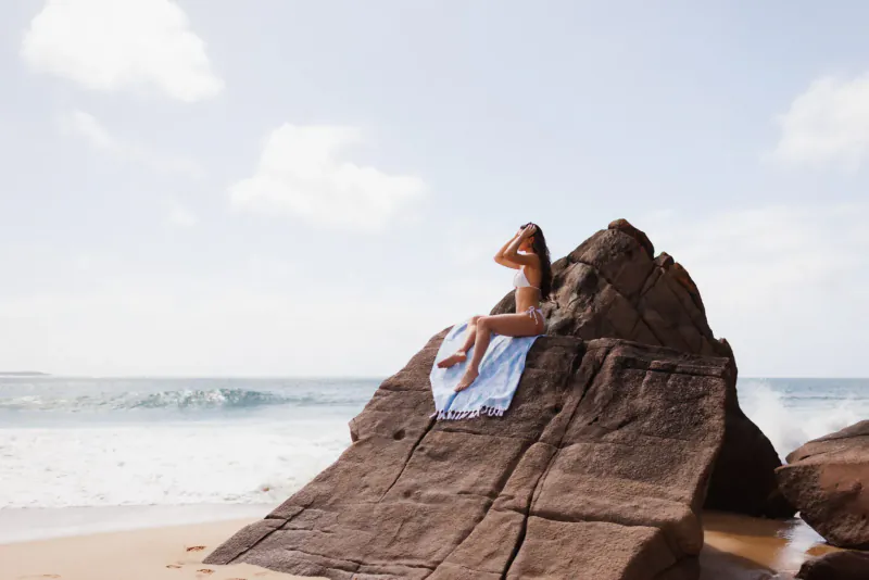 Woman in bikini sits on rocky beach outcrop with white towel, ocean waves and sky behind her