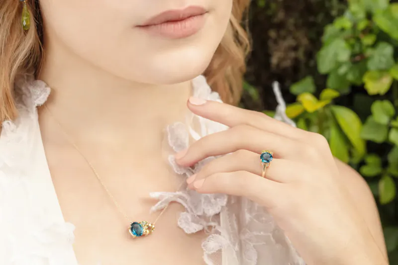 Close-up of woman in lace blouse wearing gold necklace and ring with teardrop teal gems, hand near lips, green foliage backdrop
