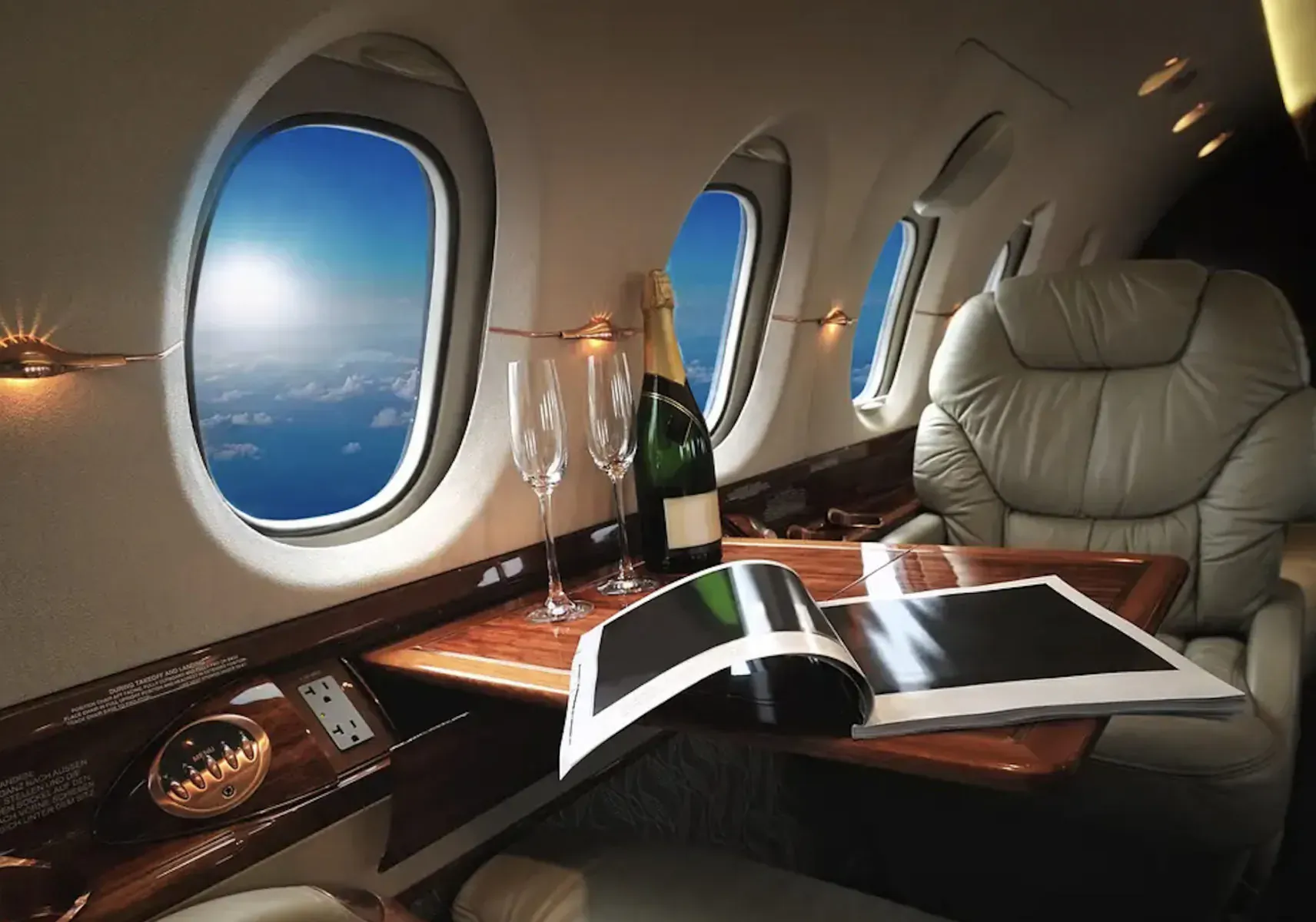 Luxury private jet interior with champagne bottle, glasses, open magazine on table, and ocean view through windows.