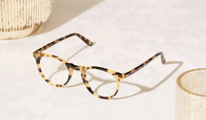 Tortoiseshell eyeglasses displayed on white surface with textured beige vase and glass in background