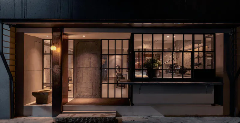 Stylish contemporary restaurant entrance at night with glass doors, warm lighting, plants, and modern interior visible.