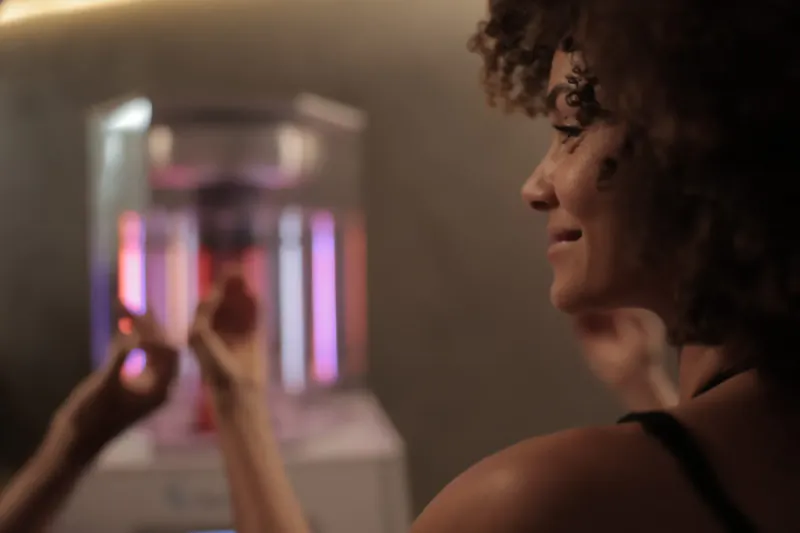 Profile of smiling Black woman with curly hair touching glowing light therapy pod in luxury wellness gallery