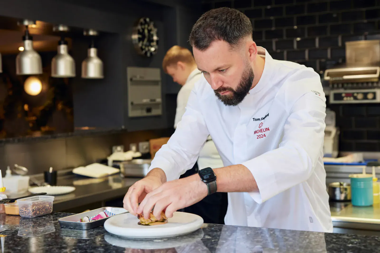 Chef Tom Shepherd in white uniform with red 'Tom Shepherd' logo, assembling a burger on a plate in modern kitchen at Wembley Stadium.