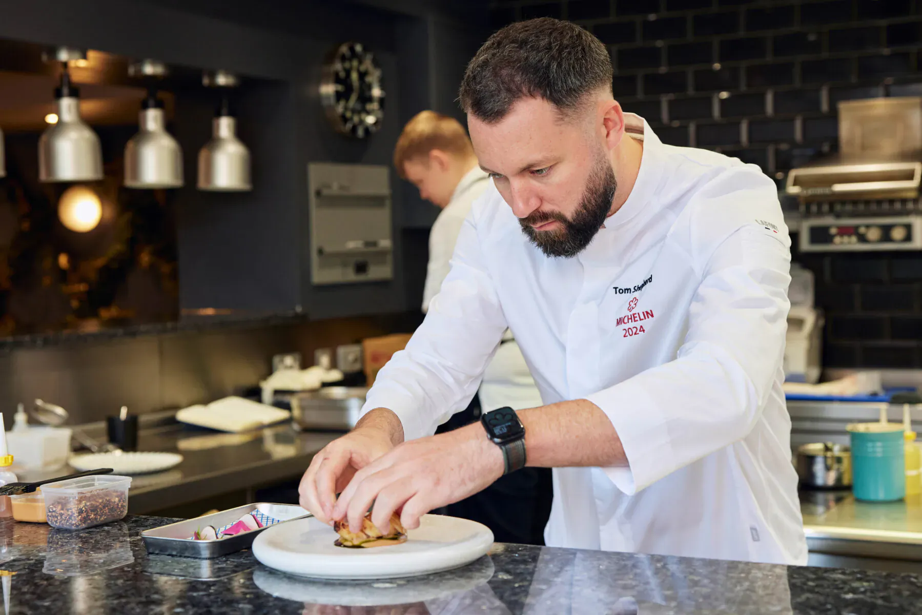 Chef Tom Shepherd in white uniform with red 'Tom Shepherd' logo, assembling a burger on a plate in modern kitchen at Wembley Stadium.