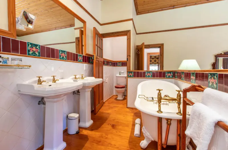 Elegant vintage bathroom at Evergreen Manor with clawfoot tub, double pedestal sinks, tiled walls, wooden floors.