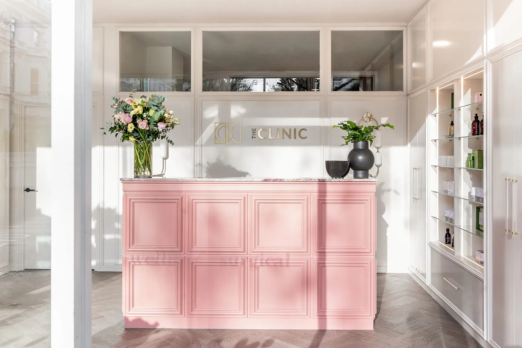 Pink reception desk at The Clinic Holland Park with yellow flowers, plants, and product shelves in modern white interior