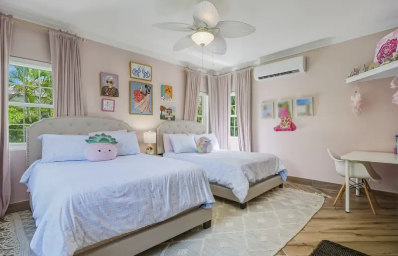 Pink girls' bedroom with two white beds, strawberry pillows, art on walls, desk, ceiling fans, and window views.