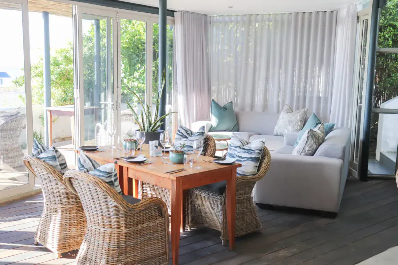 Bright sunlit indoor seating area with wooden table, rattan chairs, white sofa with teal pillows, large windows to green views.