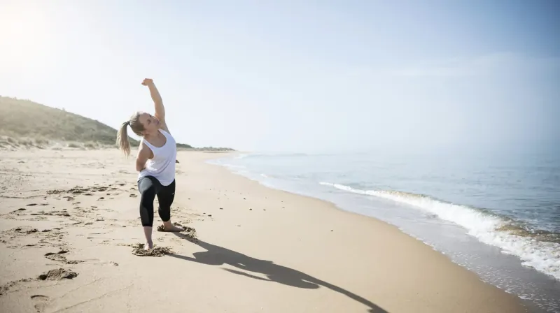 Woman in white tank top and leggings performs yoga pose with arms raised on sunny Sicilian beach