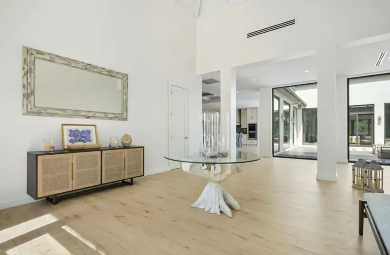 Spacious modern living room in Lyford Cay luxury home with glass dining table, wood credenza, mirror, and kitchen view