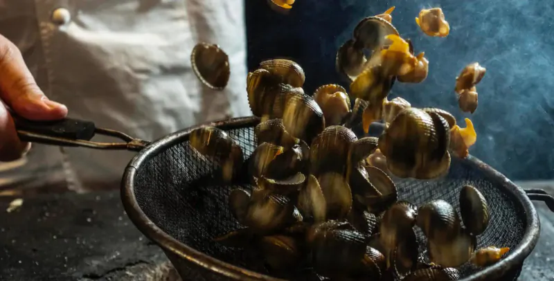 Chef's hand using tongs to toss glossy snails in a sizzling metal strainer over dark stovetop.