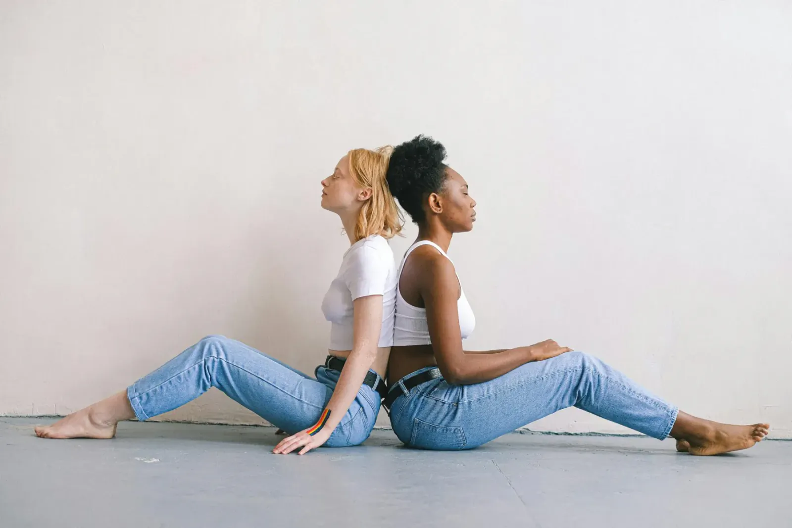Blonde and Black woman back-to-back on floor, wearing white tops and skinny jeans, legs extended.