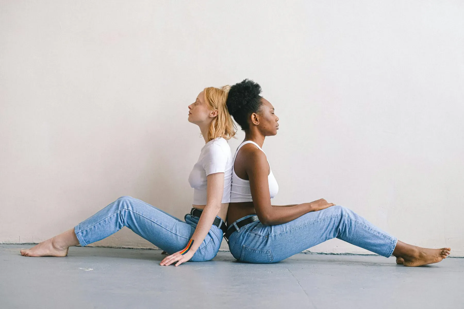 Blonde and Black woman back-to-back on floor, wearing white tops and skinny jeans, legs extended.