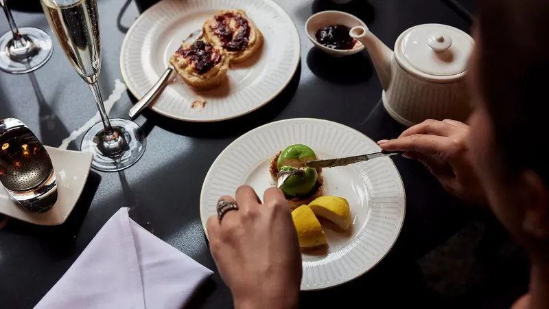 Close-up of elegant hotel table: hand cutting green dessert with mango slices, crumpets, tea pot, champagne flutes.