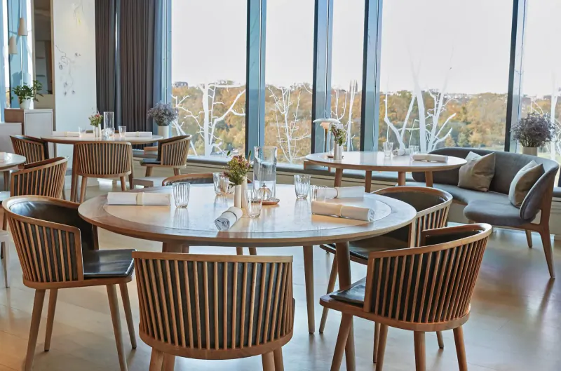 Geranium Restaurant interior: round wooden tables with white linens, flowers, and chairs by large windows with autumn trees.