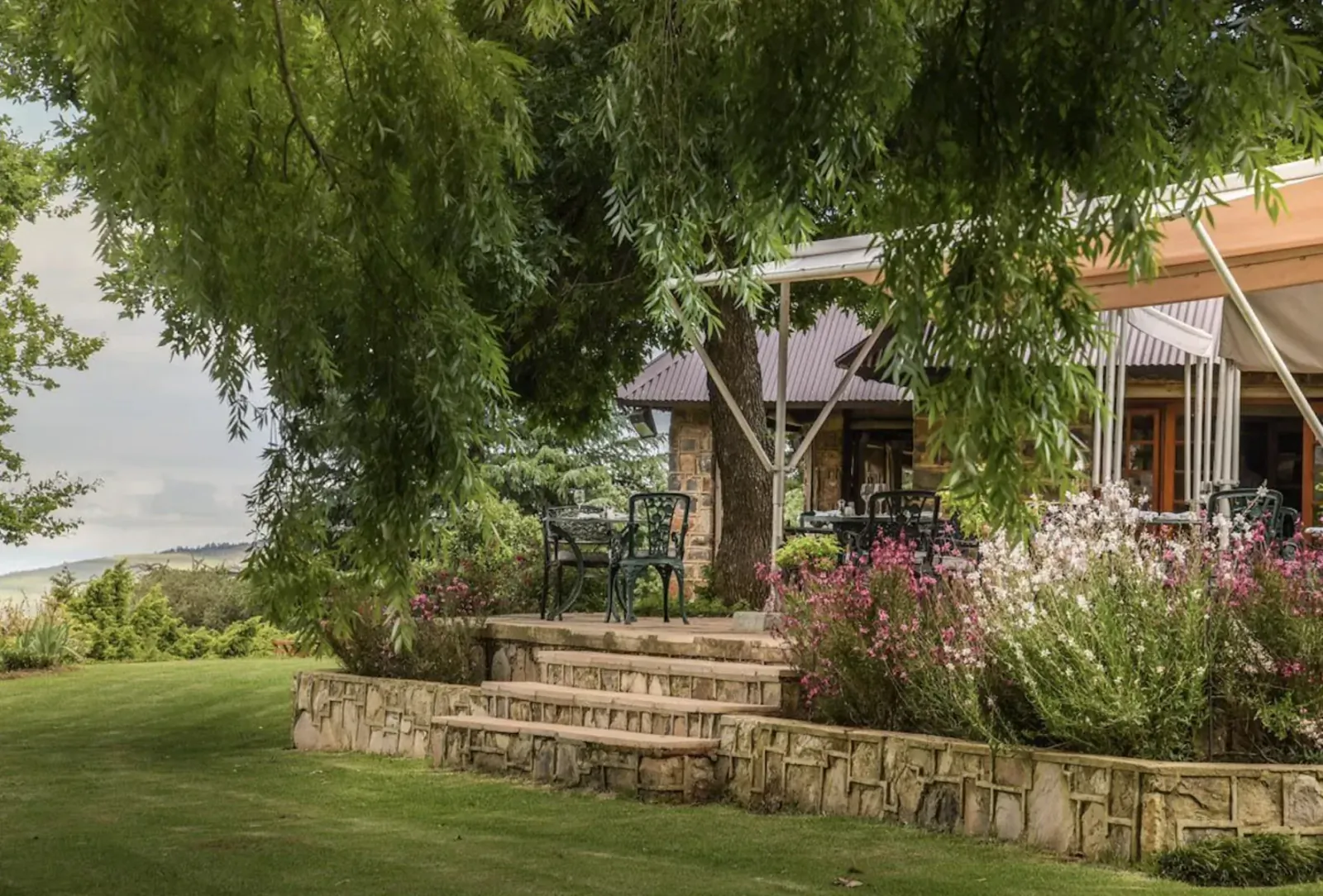 Outdoor patio restaurant with stone steps, green chairs, flowers, shaded by trees, overlooking green hills