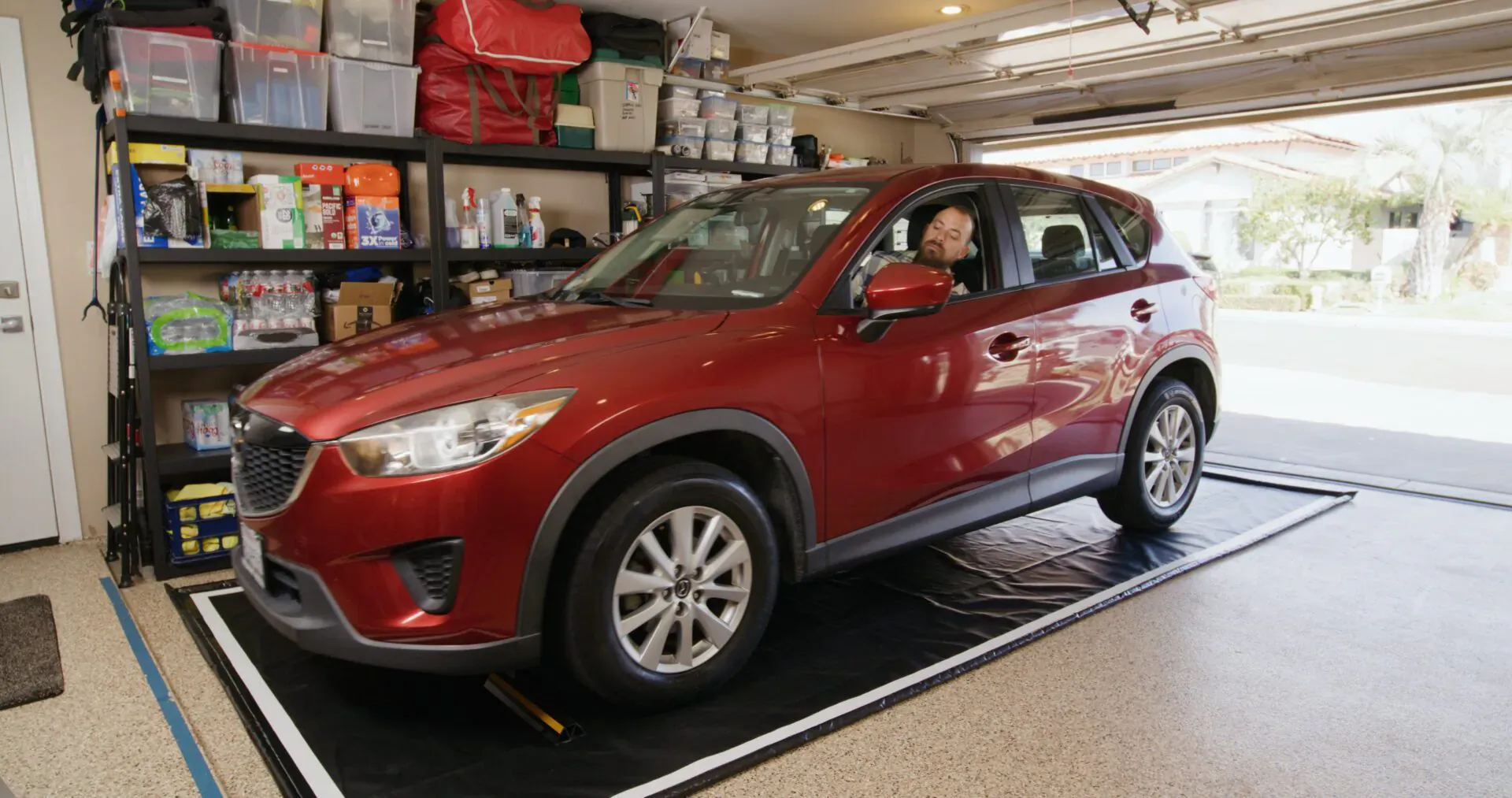Red Mazda CX-5 on black GaraMat in open garage with storage shelves, driver seated inside.