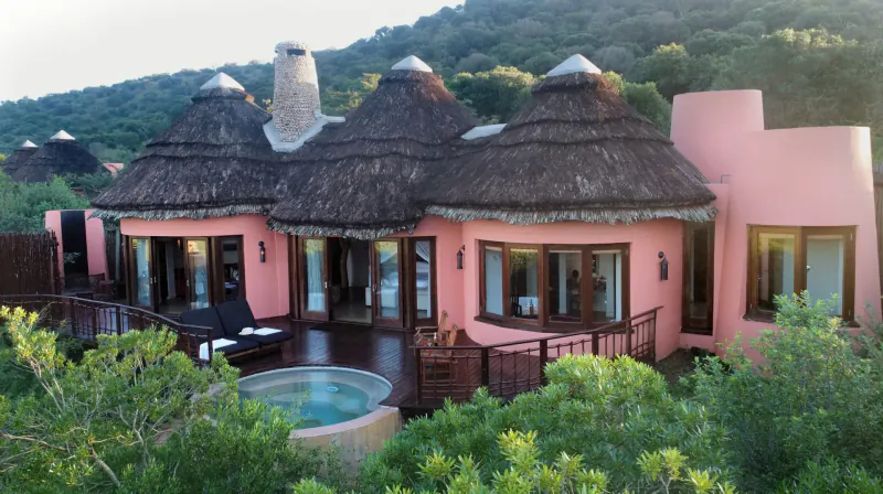 Aerial view of pink thatched-roof luxury safari villas with hot tub on wooden deck, nestled in lush green hillside vegetation.