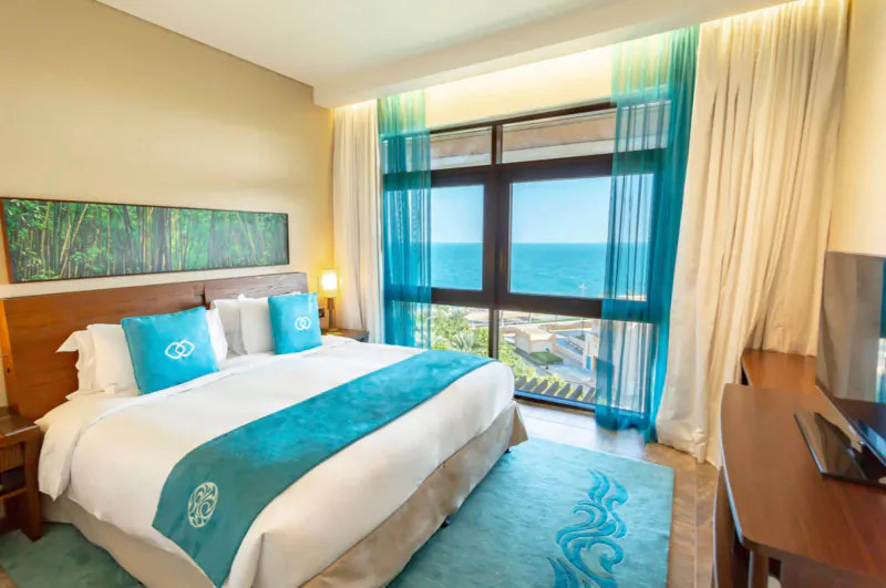 Luxurious hotel suite with king bed, teal accents, sheer curtains, and panoramic ocean view from large windows.