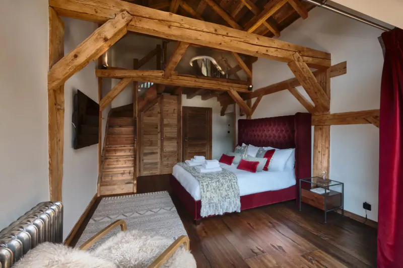 Luxurious mountainside chalet bedroom with wooden beams, red velvet bed, TV, and fur chair.