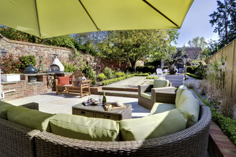 Luxury garden patio at Bray Cottages with green outdoor sofa, coffee table, built-in BBQ, and lush brick walls.