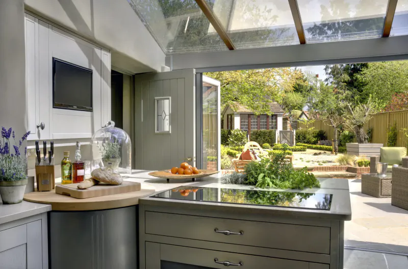 Modern luxury kitchen with glass roof, induction hob, herbs, fruits, and garden view through open doors.