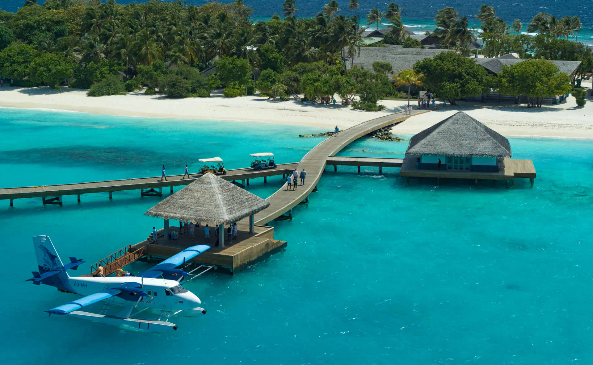 Aerial view of a blue seaplane docked at a thatched-roof pier on a white-sand beach with turquoise lagoon and palm trees in Maldives.