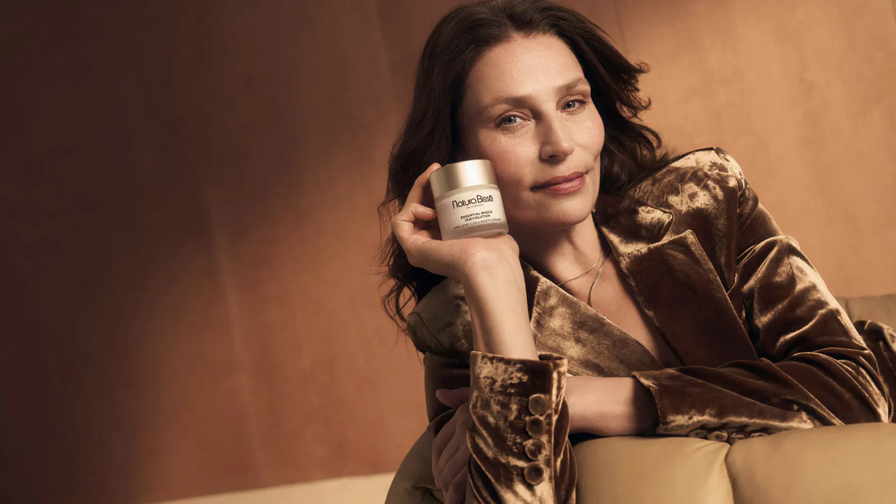 Woman in bronze velvet blazer holding silver skincare jar, smiling on warm background