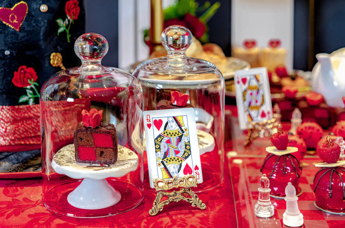 Elegant afternoon tea display with Queen of Hearts cakes under glass domes, red roses, strawberries, and chess pieces on red table.