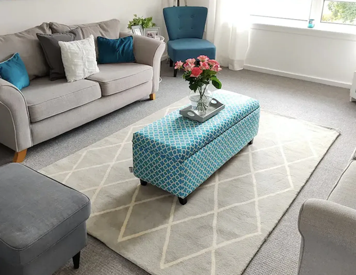 Cozy living room with gray sofa, turquoise ottoman holding pink roses in glass vase, on cream rug.