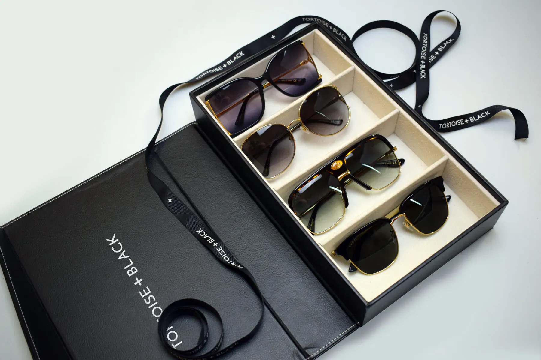 Open black Tortoise+Black luxury eyewear box displaying six pairs of stylish sunglasses in compartments with satin ribbons.