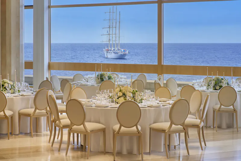 Elegant round table set with white chairs and flowers in Fairmont Monte Carlo ballroom, sunset sea view with tall ship through windows.